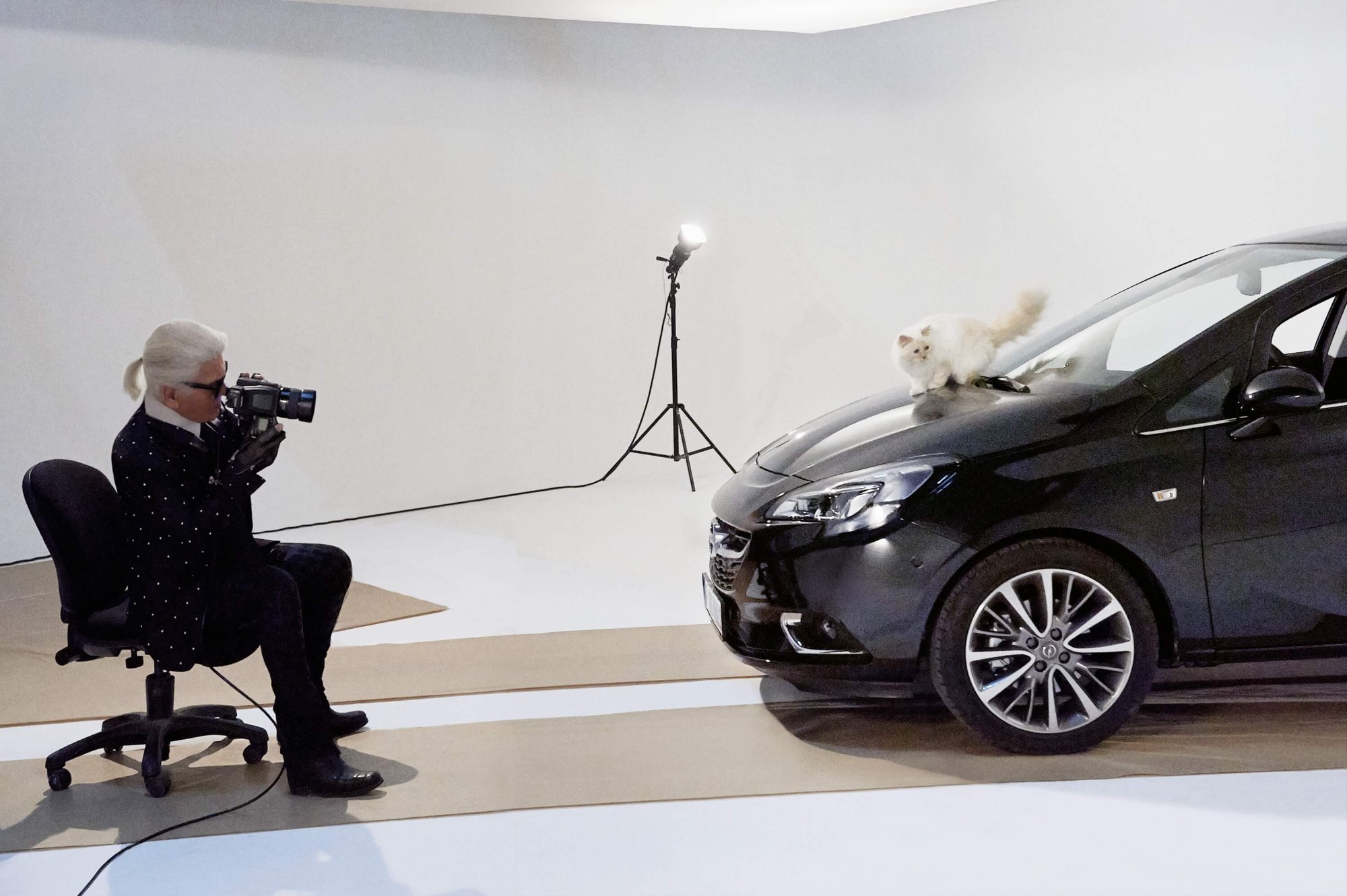 Karl Lagerfeld sitzt auf einem Bürostuhl und fotografiert seine Katze Choupette, die auf der Motorhaube eines dunklen Opel Corsa posiert – Fotostudio-Setting